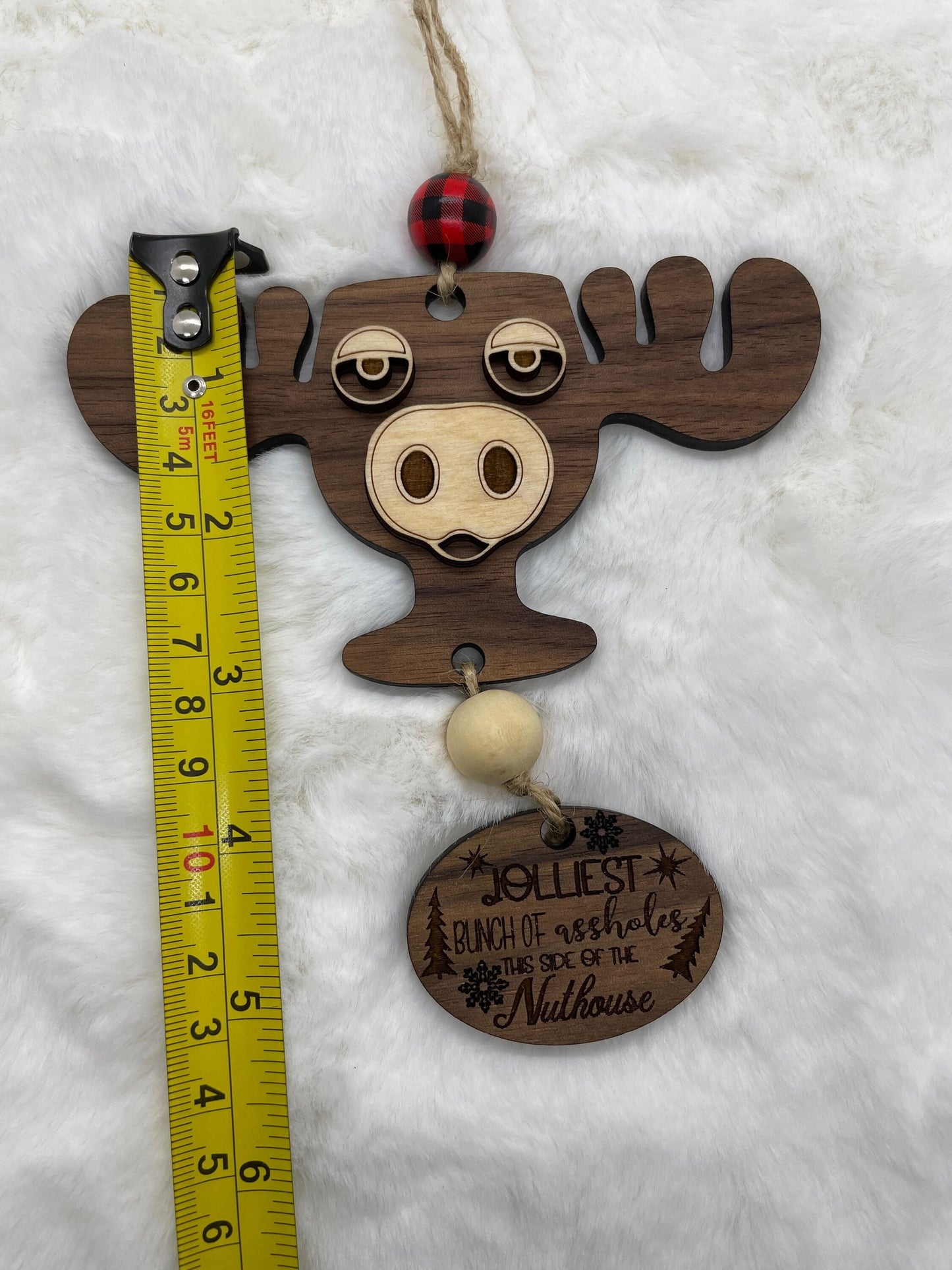 Wooden moose ornament with humorous tag and measuring tape for scale on a white background