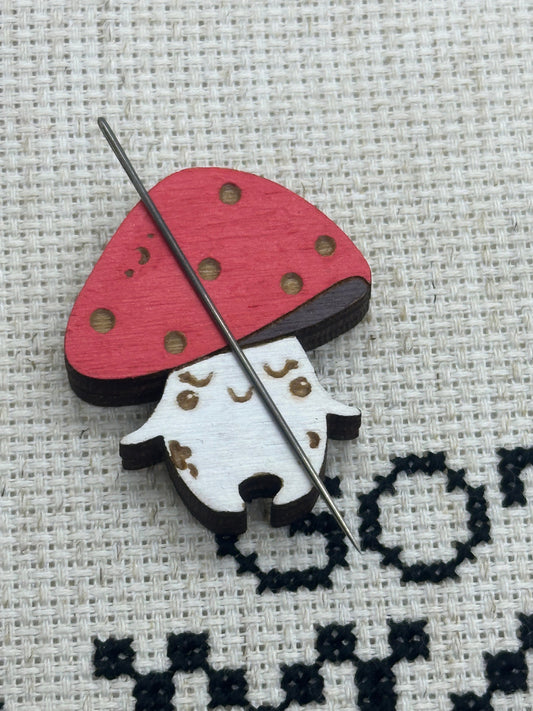 Cross-stitched design of a mushroom with a red hat on fabric with a needle threaded through the fabric.