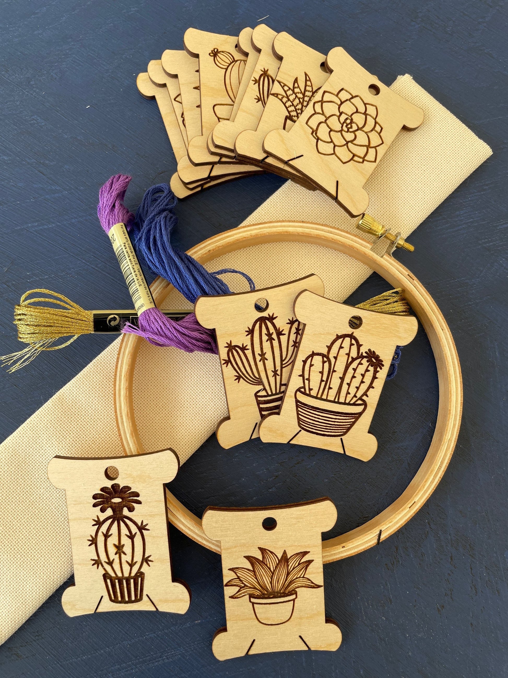 Wooden tags with cactus designs, embroidery hoop, and yarn on a blue background