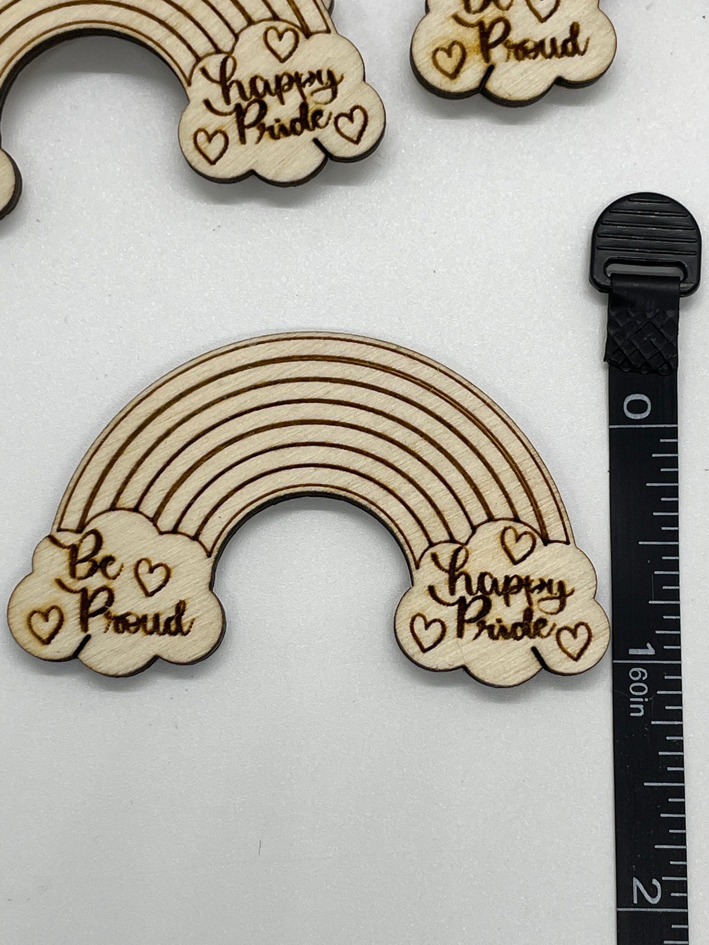 Wooden rainbow-shaped decorations with 'Be Proud' and 'Happy Pride' text on a white background with a ruler for scale.