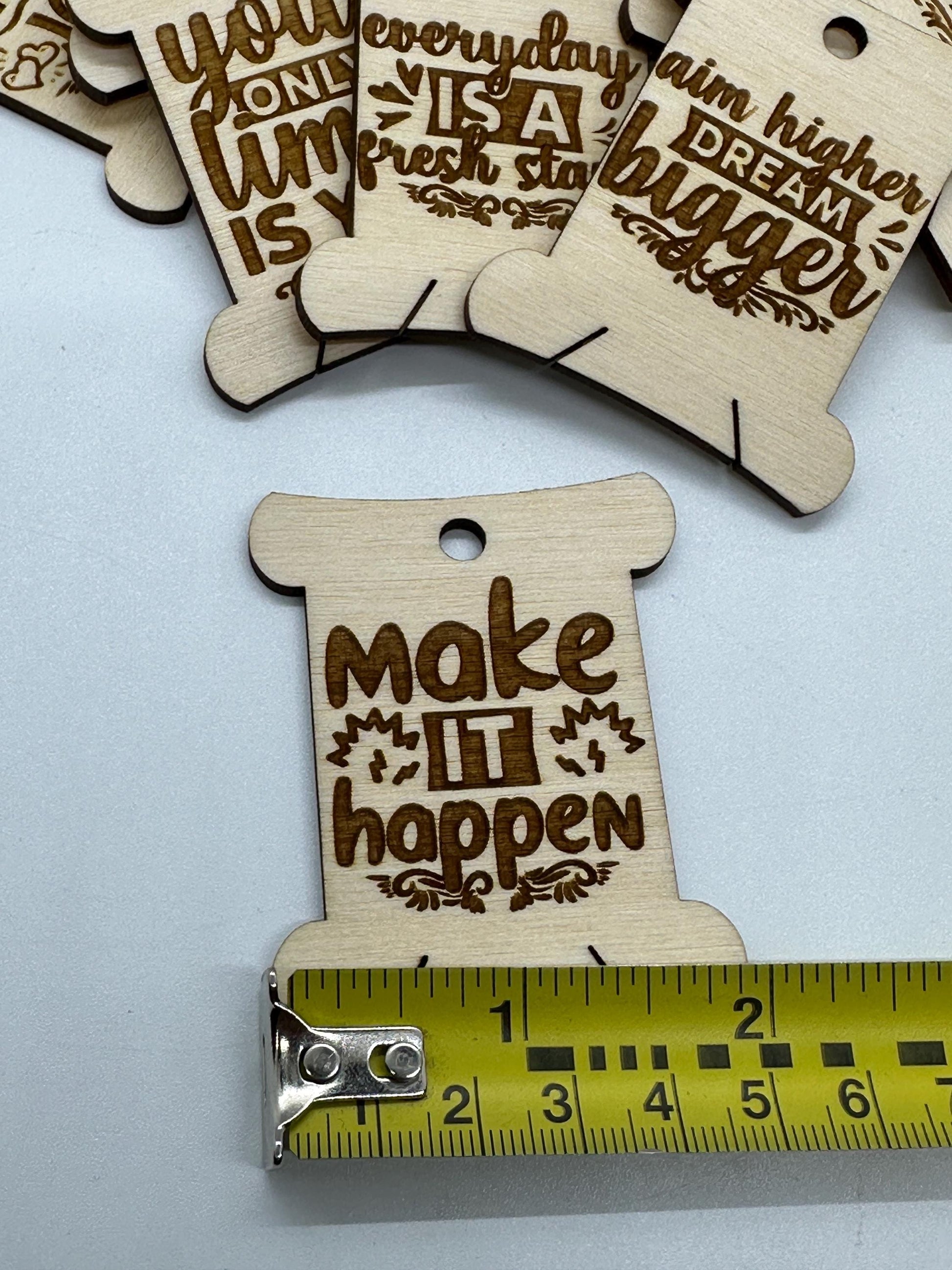 Wooden motivational tags with engraved text on a white background, one tag is measured by a ruler.