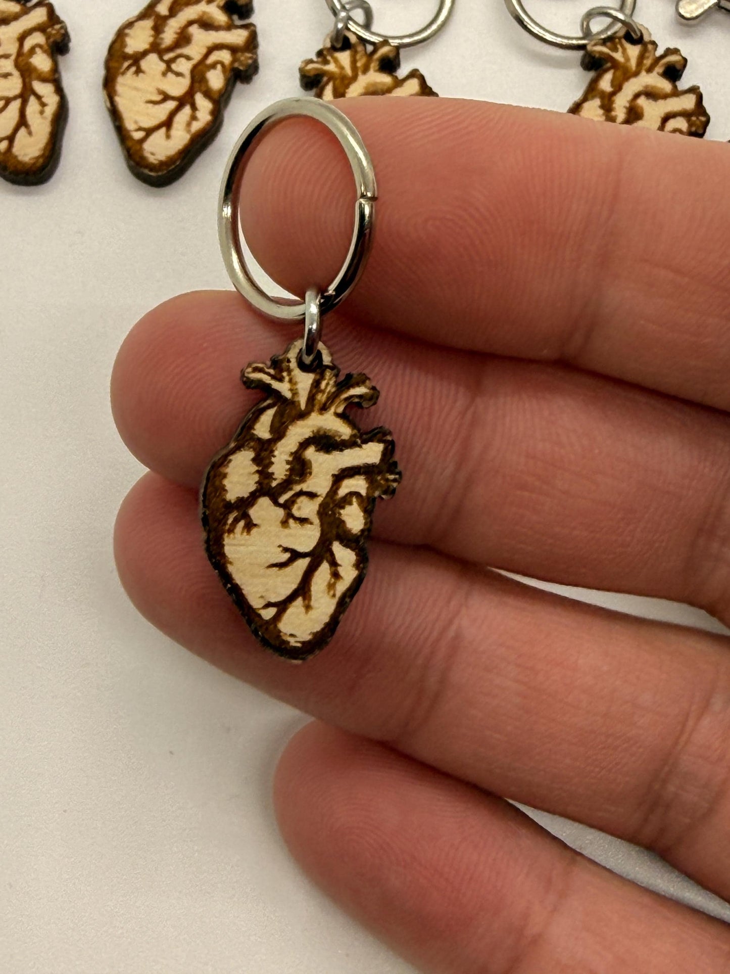 Wooden anatomical heart keychain held between fingers with blurred anatomical heart models in the background.